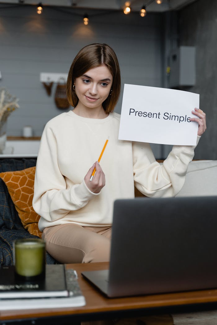 about-img Woman teaching English online holding Present Simple flashcard, engaging in video call.