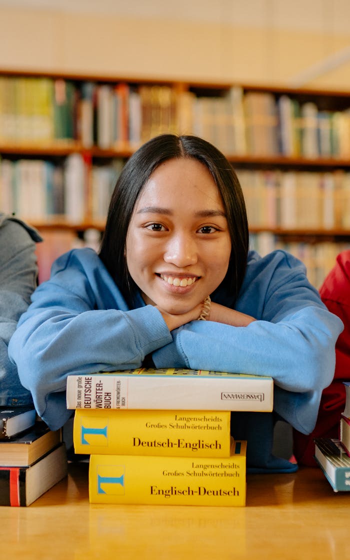 gallery-05 Smiling student in a library leaning on a stack of books, symbolizing education and learning.